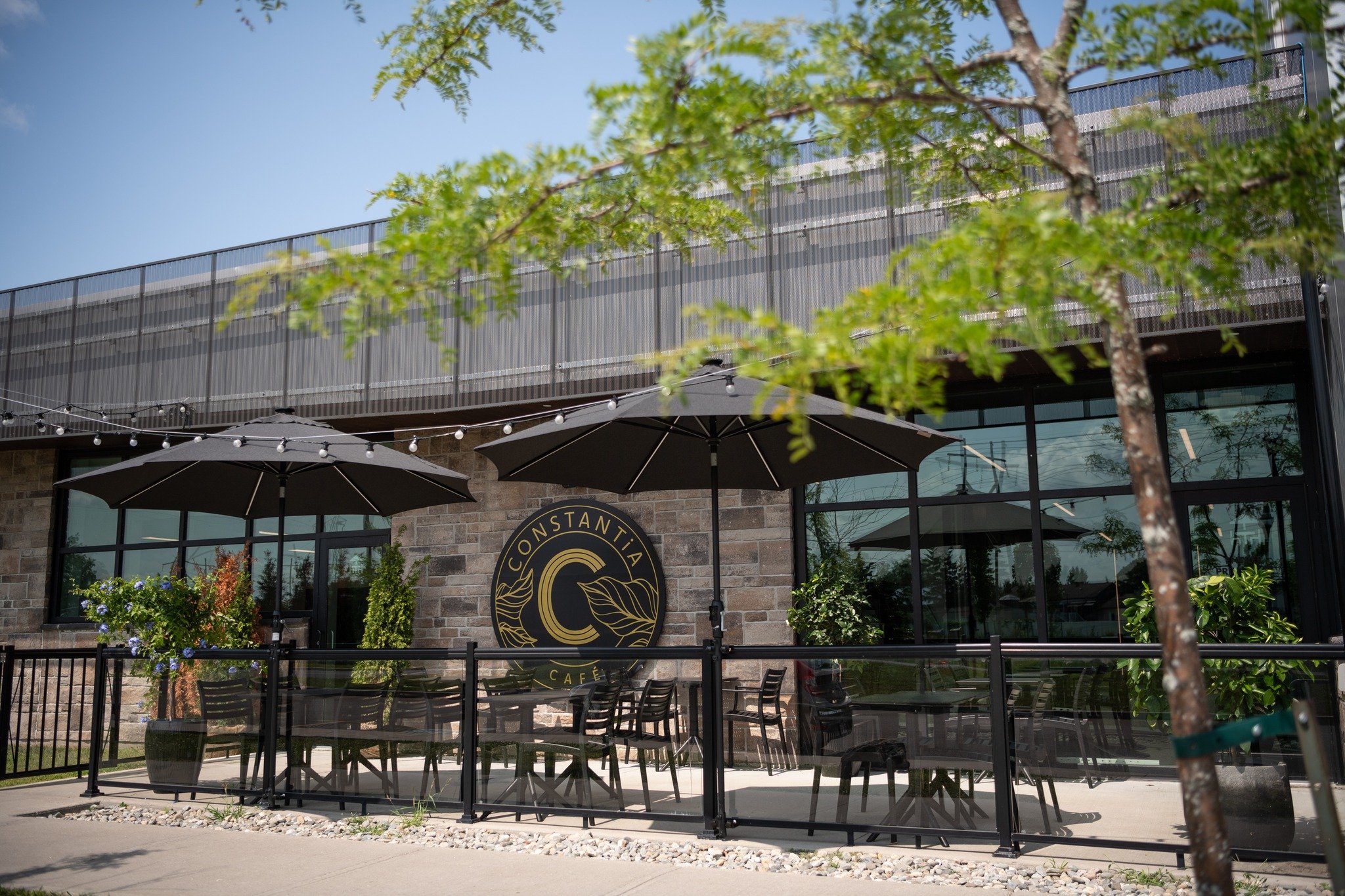 Terrasse ensoleillée du Café Constantia à Saint-Constant — parasols noirs et logo mural doré